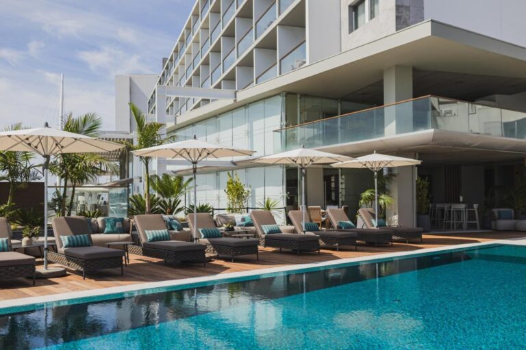 Modern hotel pool area with blue-tiled pool, lined with cushioned lounge chairs and umbrellas on a wooden deck. Large white building in the background features multiple balconies, glass railings, and tall windows. Lush greenery adds to the serene ambiance.