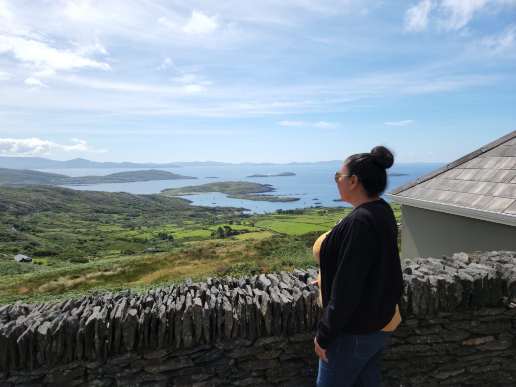 A woman stands beside a stone wall, gazing at a scenic view of lush green hills and a vast body of water with small islands. The sky above is mostly clear with scattered clouds. Dressed in casual clothes with her hair in a bun, she reflects on the best deals life can offer amidst such beauty.