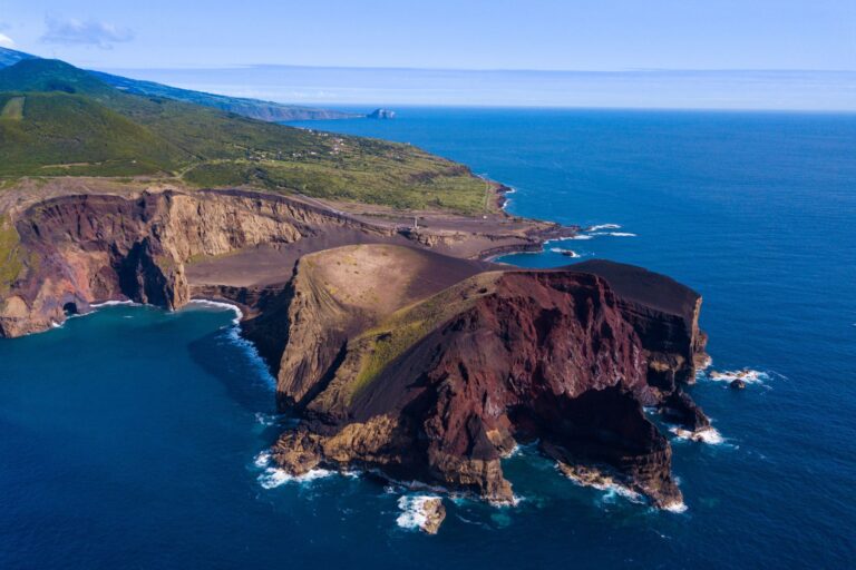 Aerial view of a rugged coastline featuring dramatic cliffs and rocky formations jutting into the blue sea. The landscape is a mix of verdant green hills and barren brown volcanic terrain, with waves crashing against the rocky shores.