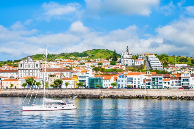 A vibrant coastal town featuring colorful buildings and hillside greenery. A white sailboat floats on calm blue waters in the foreground. The town includes various architectural styles, with a notable church and lush green hills in the background under a partly cloudy sky.