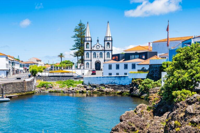 A scenic coastal town features a white church with two tall steeples, surrounded by colorful, traditional houses with red-tiled roofs. The foreground showcases a calm waterway with rocky shores, greenery, and a clear blue sky overhead.