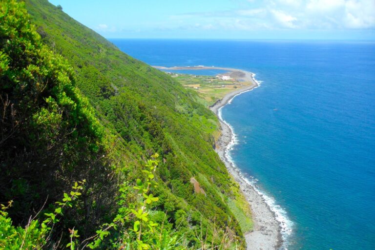 A coastal view features a lush green hillside descending to a narrow, rocky shoreline that borders a vibrant blue sea. In the distance, a flat area with sparse vegetation extends out into the ocean under a bright blue sky with scattered clouds.