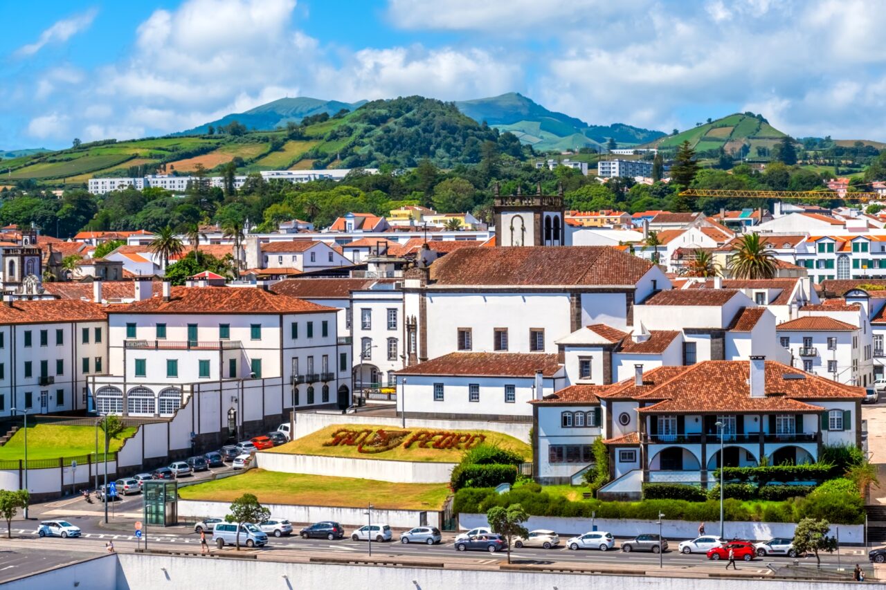 Scenic view of a town with white buildings and terracotta rooftops against a backdrop of green hills and blue sky. A church tower and a sign reading "São Pedro" are visible. A line of cars is parked along the street in the foreground.