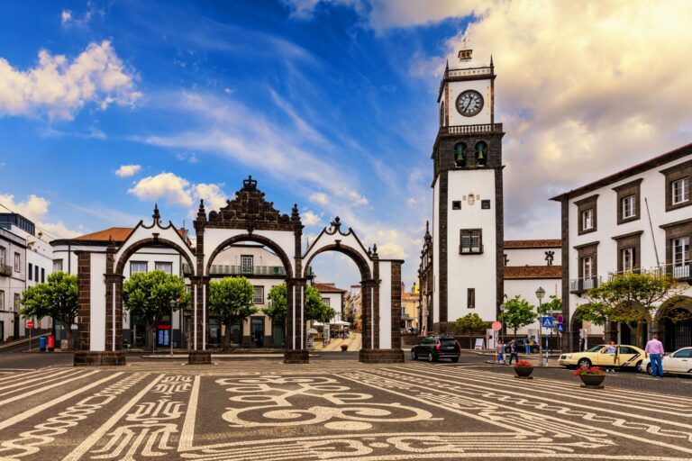 A picturesque town square features three arches in front of a historical building and a white clock tower with a black roof. The ground is adorned with an intricate black and white patterned mosaic, surrounded by trees, parked cars, and people strolling.