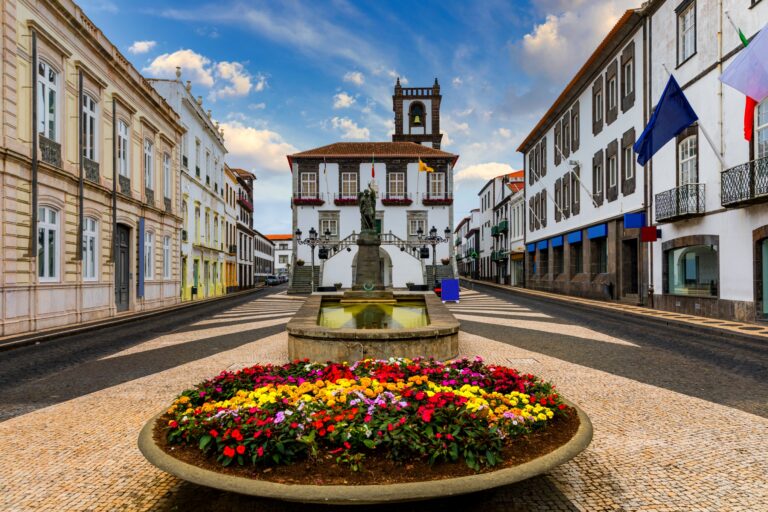 A vibrant town square with a central fountain surrounded by colorful flowers. Lined with classical buildings on either side, the scene leads to a prominent, historic clock tower at the end of the street under a bright, partly cloudy sky.