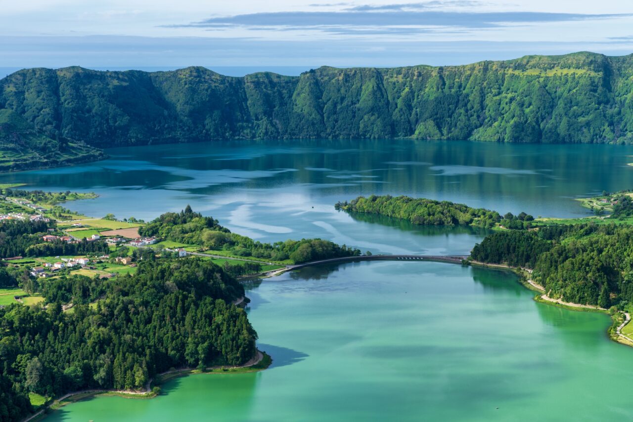 A picturesque landscape of a strikingly blue and green dual-colored lake, surrounded by lush green hills and forests. A small bridge connects two sections of the lake, with a quaint village visible on the left side, nestled among the greenery.