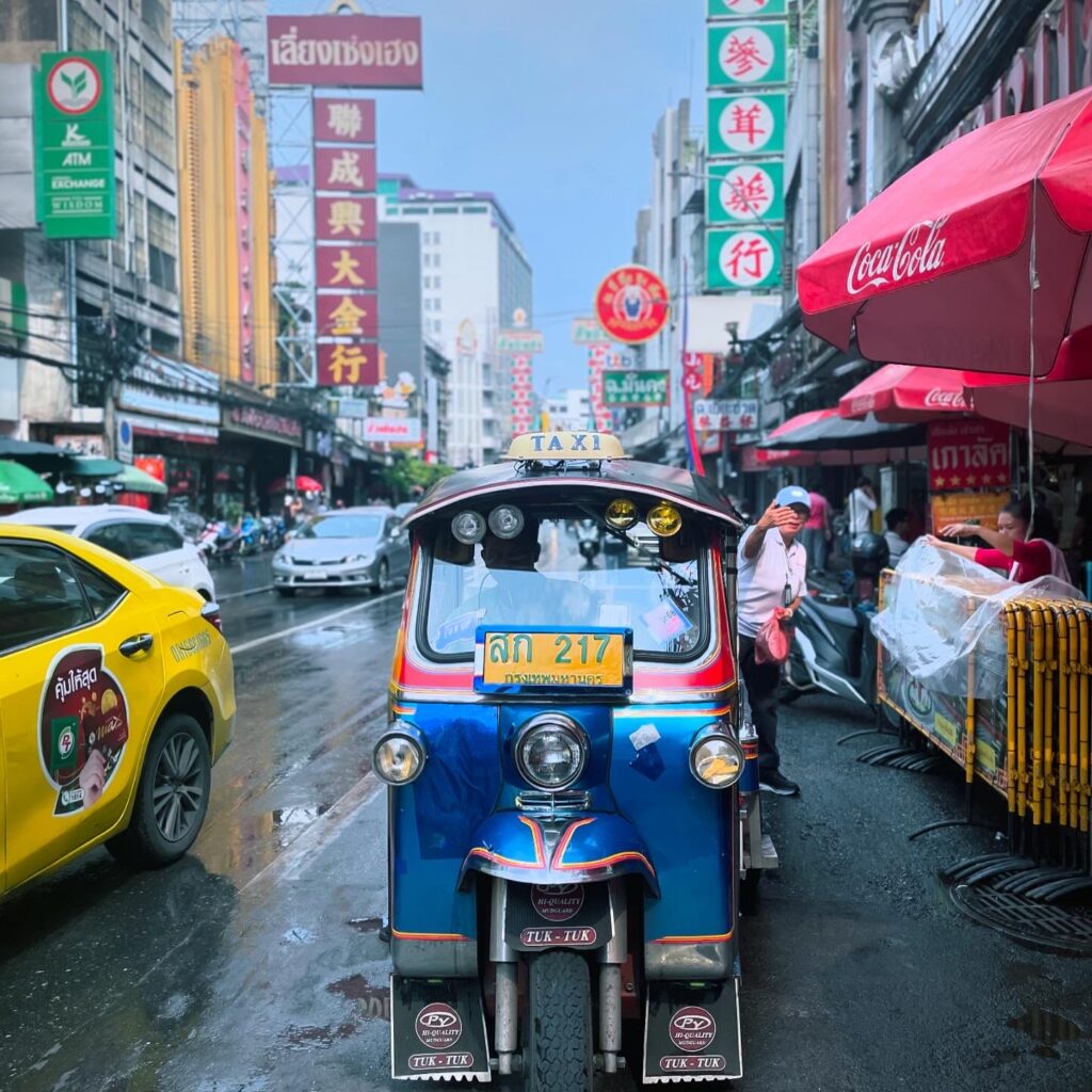 A blue tuk-tuk is parked on a wet street in a bustling city with vibrant signs in various languages, reminiscent of varied wine regions. A yellow taxi is nearby, and people stroll under red umbrellas. The atmosphere is lively and colorful.