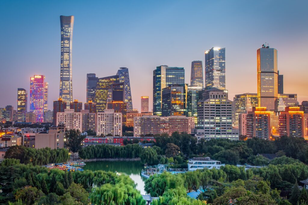 A vibrant cityscape of Beijing at dusk with illuminated skyscrapers, including the distinctive China Zun Tower, and a lush green park in the foreground under a clear sky.