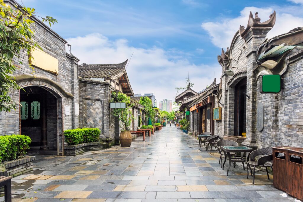 A picturesque street in an ancient Chinese town featuring traditional gray brick buildings with ornate roofs. The walkway is lined with greenery and outdoor seating, leading to a distant view of modern city skyscrapers under a blue sky.