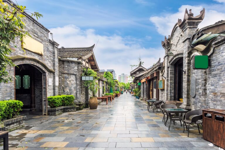 A picturesque street in an ancient Chinese town featuring traditional gray brick buildings with ornate roofs. The walkway is lined with greenery and outdoor seating, leading to a distant view of modern city skyscrapers under a blue sky.