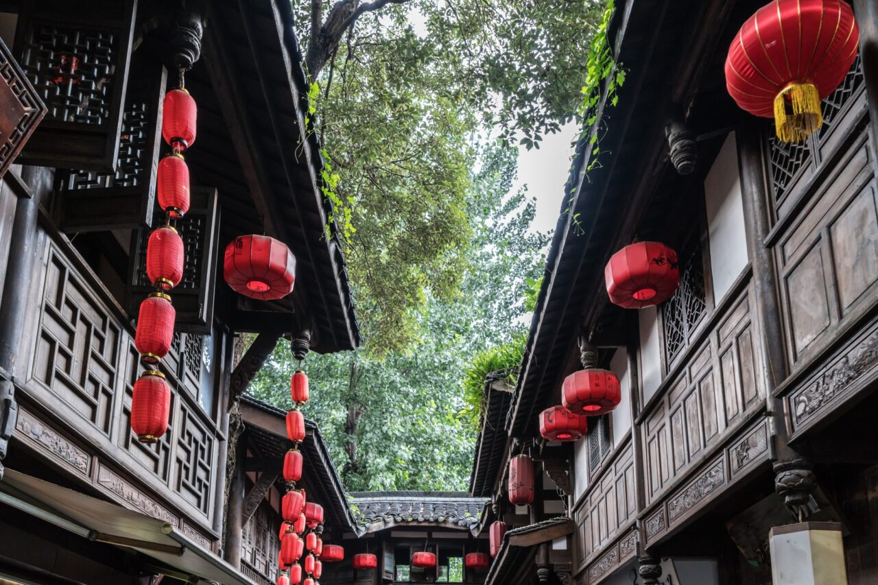 A narrow alley between traditional wooden buildings adorned with red lanterns. The structures feature intricate woodwork, and lush green trees can be seen above, adding a natural backdrop to the scene.