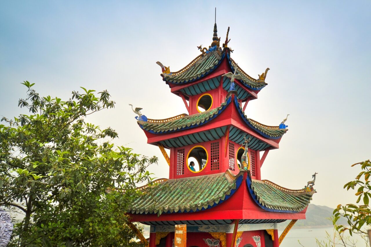 A vibrant Chinese pagoda with multiple tiers and ornate details, featuring red walls and green tiled roofs. Decorative figures adorn the roof edges. Trees surround the structure, and a hazy sky forms the background.