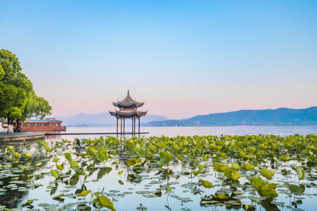 A serene lake scene featuring a traditional Asian pavilion on stilts over the water, surrounded by green lotus leaves. Trees line the left shore, and distant mountains under a clear blue sky complete the tranquil landscape.