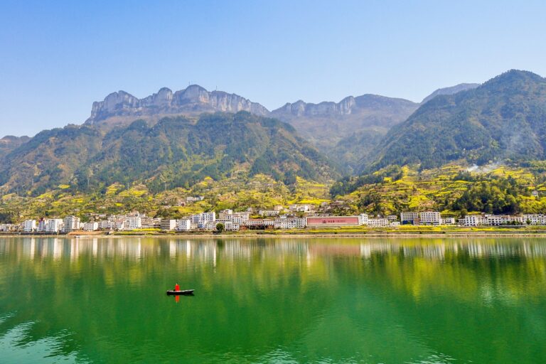 A serene landscape featuring a calm green lake with a single person in a small boat, surrounded by a town with white buildings. The backdrop includes lush green mountains under a clear blue sky.