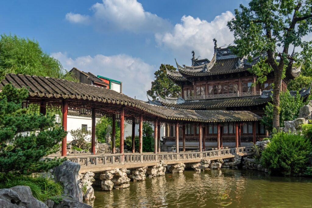 A traditional Chinese garden with a wooden bridge over a pond leads to an intricately designed building with curved roofs and ornate details. Lush greenery surrounds the tranquil scene under a partly cloudy sky.