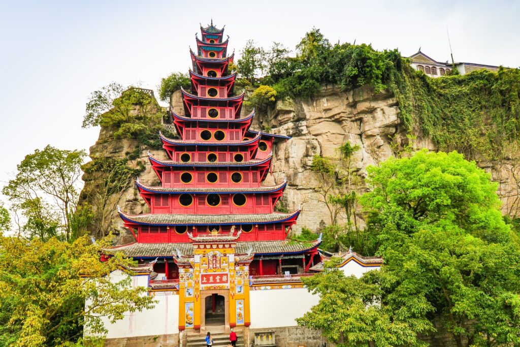 A vibrant, multi-tiered pagoda with red and black detailing is built against a rocky cliff. Lush greenery surrounds the structure, adding a natural background to the scene. A few visitors are seen near the entrance.