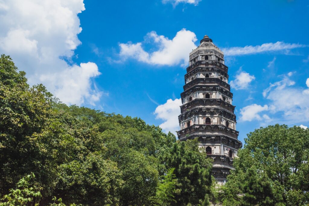 A tall, multi-tiered stone pagoda rises above lush green trees under a bright blue sky with fluffy white clouds. The structure features a series of overhanging roofs and decorative elements, showcasing traditional architecture.