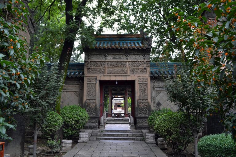 Ancient Chinese temple entrance surrounded by lush greenery. The ornate stone gate features intricate carvings, with a pathway leading through a series of doorways framed by trees and bushes.