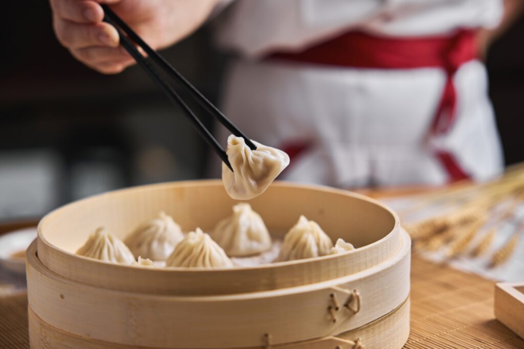 A person using chopsticks to lift a dumpling from a bamboo steamer filled with several dumplings. The person is wearing a white outfit with a red apron.