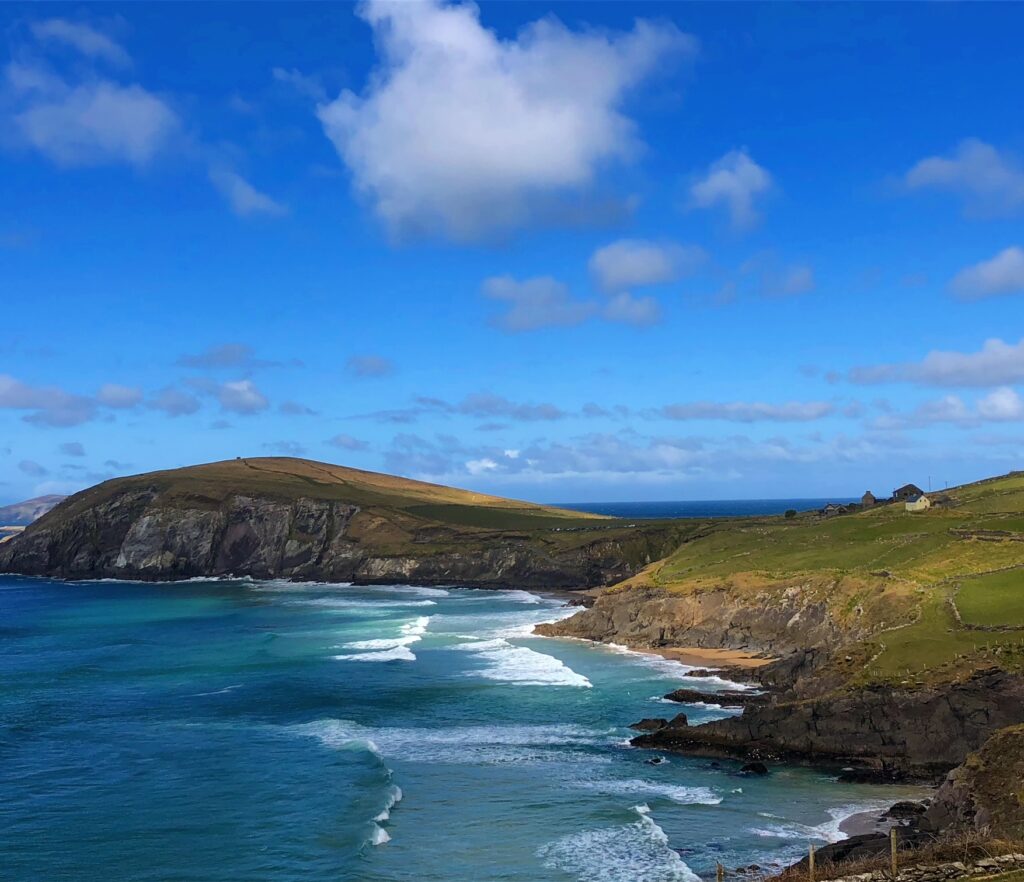 A scenic coastal view with clear blue skies, rocky cliffs, and white waves crashing against the shoreline resembles some of the world's most beautiful wine regions. The landscape features grassy hills and a few small structures near the cliffs, under a sky dotted with fluffy clouds.