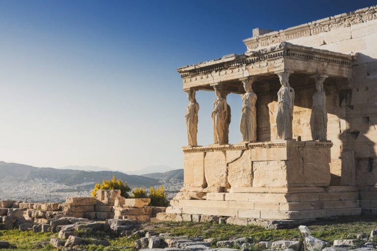 Ancient Greek temple with six sculpted female figures as columns, known as Caryatids, under a clear blue sky. The temple is situated on a rocky hillside with a distant view of a city and mountains.