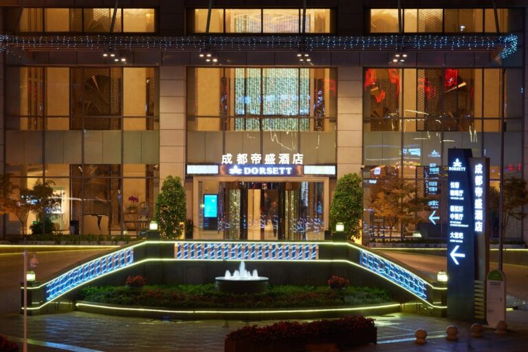 Entrance of a hotel at night with a sign that reads "Dorsett." The entrance is well-lit with decorative lights and features a small fountain in front. A lit directory sign is on the right, surrounded by greenery and illuminated steps.