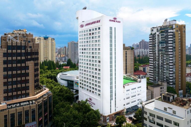 A tall, white modern hotel building labeled "Crowne Plaza" is surrounded by other city buildings and lush green trees. The sky is partly cloudy, suggesting a mixture of sun and clouds.