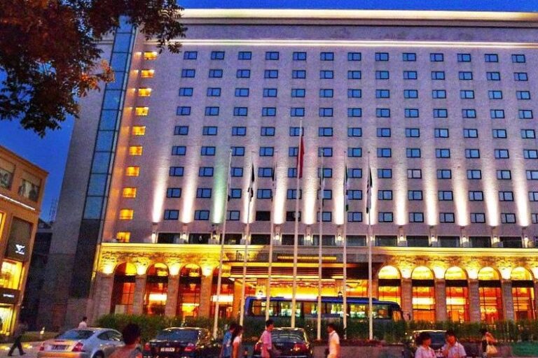 A large, illuminated hotel building at night with multiple rectangular windows arranged in a grid pattern. The entrance is brightly lit with arches and columns. Cars and people are visible in the foreground.