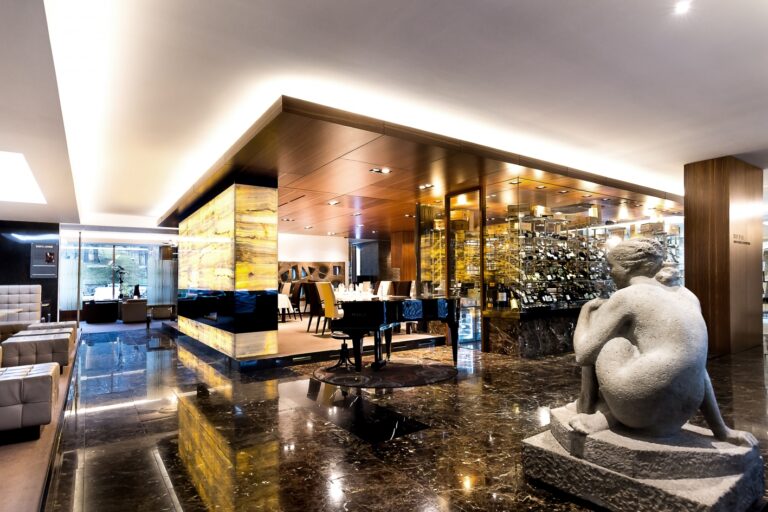 Luxurious interior of a modern hotel lobby featuring a backlit onyx wall, a glossy black grand piano, and a gray sculpture of a seated figure. The area is elegantly furnished with polished marble floors and sleek seating arrangements.