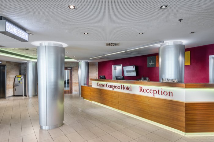 Hotel lobby with a modern design. A wooden reception desk with "Clarion Congress Hotel Reception" is in the center. Silver columns and a red wall with screens are in the background. A sign labeled "Congress" hangs from the ceiling.