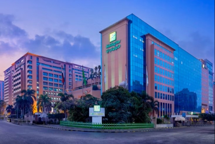 A large, modern hotel building with a glass facade and Holiday Inn signage. It's surrounded by palm trees under a blue and pink sky at dusk.