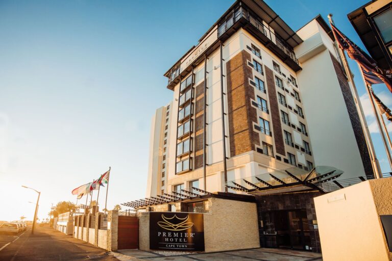 Exterior view of a tall hotel building with a modern design, featuring multiple floors and large windows. The signage reads "Premier Hotel Cape Town." Several flags are flying near the entrance, and there is a clear blue sky in the background.