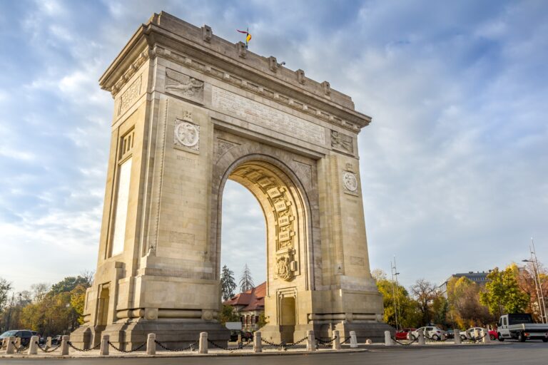 The image shows a large, historic triumphal arch with detailed carvings, set against a blue sky. The structure is surrounded by a paved area and trees with autumn foliage.