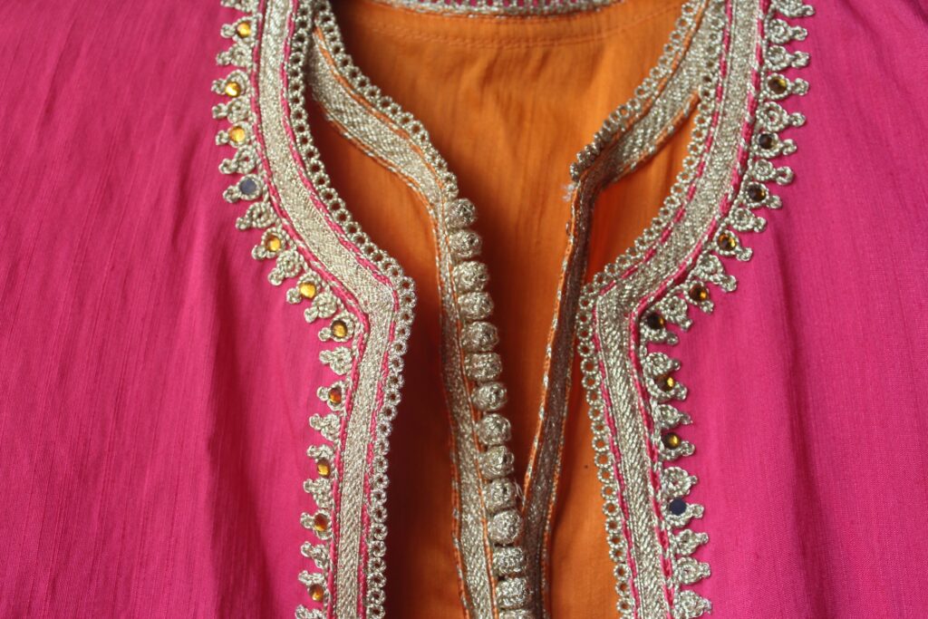 Close-up of a vibrant pink and orange traditional garment with intricate gold embroidery and beadwork along the neckline. The embroidery features ornate patterns and decorative details.