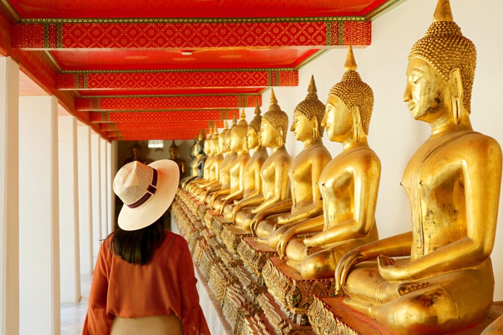 A person wearing a hat and orange clothing observes a row of golden Buddha statues inside a temple with a red and gold patterned ceiling.