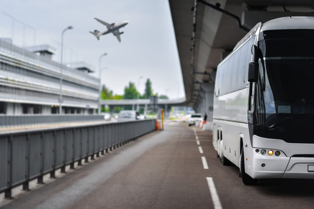 A white tour bus is parked on a road under an airport terminal bridge, highlighting the convenience of free airport transfers. In the background, a plane is taking off, capturing the essence of travel and transit at a bustling airport.