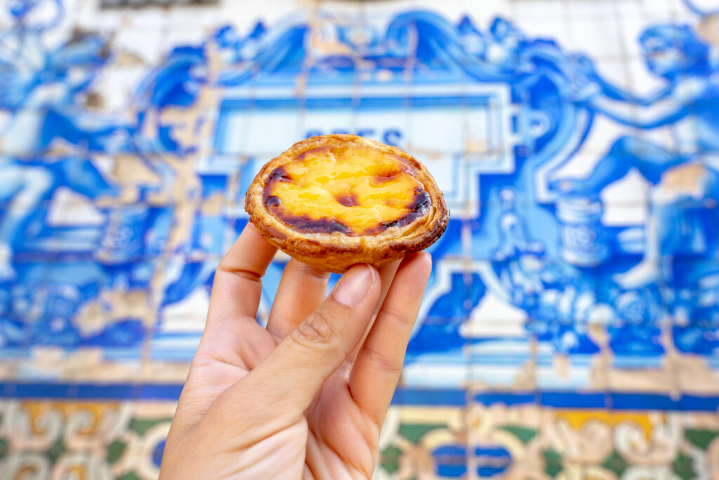 A person holds a Portuguese custard tart, a delightful example of Portuguese food, with its golden crust and creamy center set against a backdrop of blue and white traditional tile art.