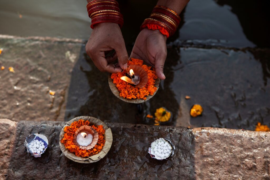 Hands placing a lit diya surrounded by orange marigold petals on water steps, alongside other small floral arrangements and floating candles. Ritualistic setting suggests a cultural or religious ceremony.