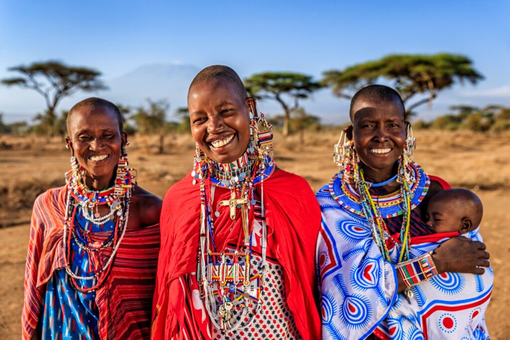 Three smiling people in traditional Maasai attire, adorned with colorful beads and earrings, stand in a sunlit, open landscape with acacia trees. One carries a baby in a patterned wrap. The backdrop features distant mountains and a clear blue sky.