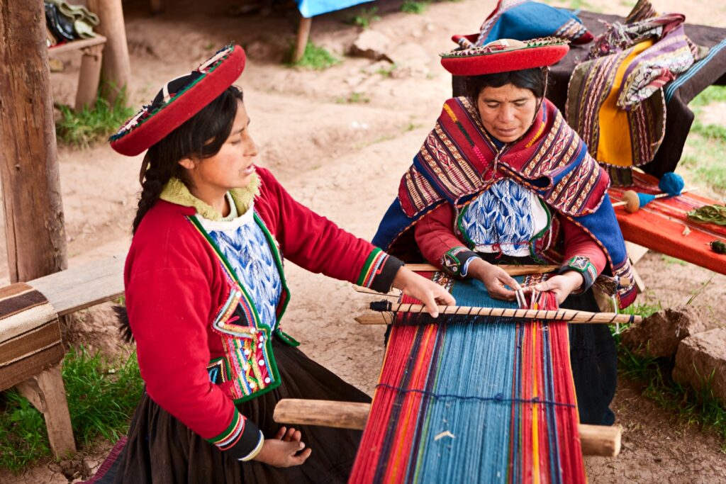 Two women in traditional Andean clothing work on weaving a colorful textile on a wooden loom. The women are seated on the ground, focusing on the intricate design, with earthy surroundings and woven fabrics nearby.