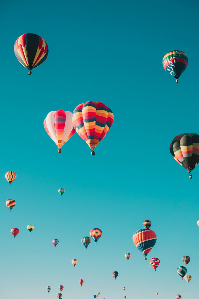 A sky filled with colorful hot air balloons of various patterns and sizes floating under a clear blue sky. The vibrant colors create a striking contrast against the backdrop.