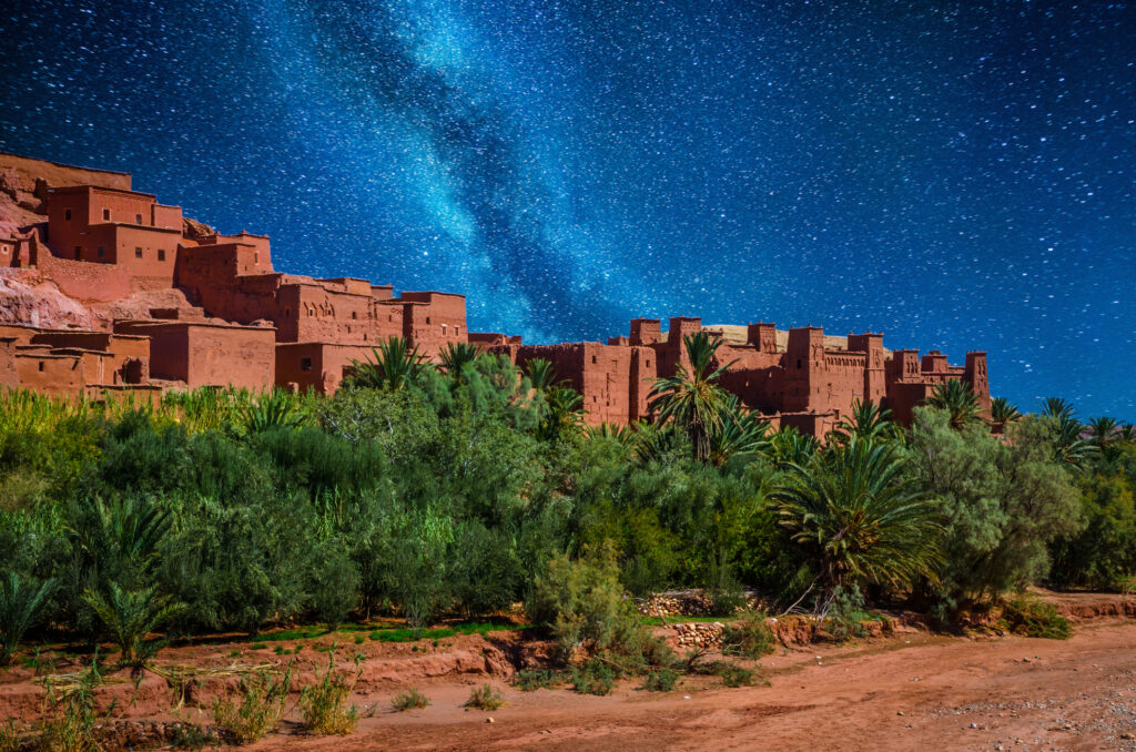 Ancient clay buildings under a starry night sky with the Milky Way visible offer a glimpse into timeless beauty. Surrounded by lush greenery, these structures provide an enchanting contrast to the clear, vibrant blue night—a perfect setting for discovering hidden deals in nature's embrace.