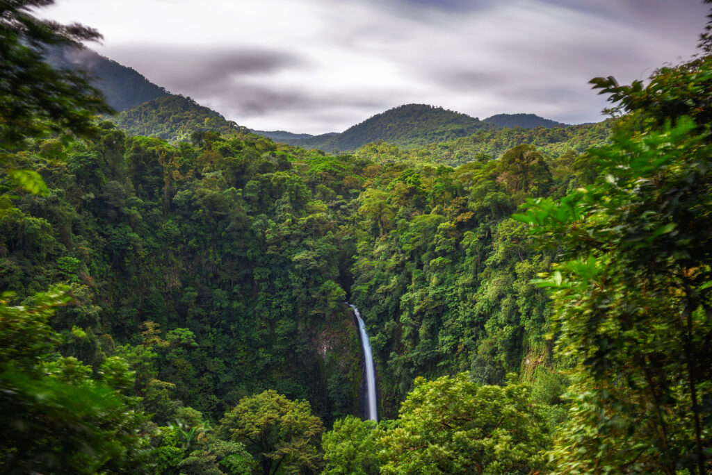 A waterfall cascades into a lush rainforest valley surrounded by dense foliage and distant mountains under a cloudy sky. This tropical paradise exudes serene beauty while embracing the Sustainability Pledge for responsible tourism through Smart Impact Travel initiatives.