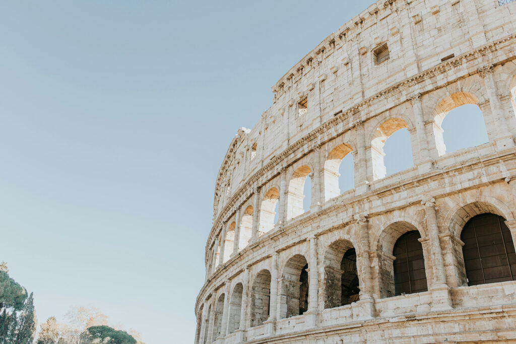 The image shows an upward view of the ancient Colosseum in Rome against a clear blue sky. The sun casts a gentle light on the stone arches, highlighting its historic structure. Trees are partially visible at the edge.