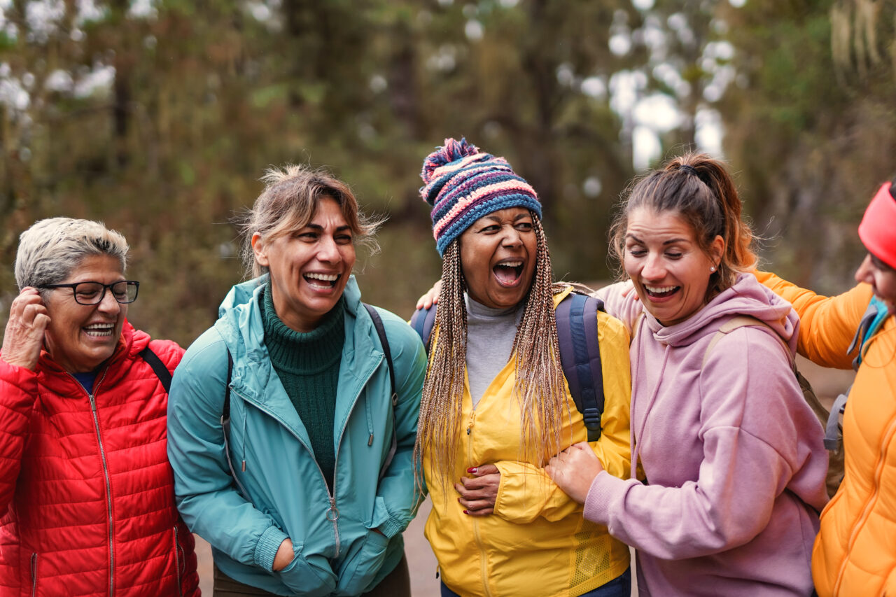 A group of five women laughing together in a forest. They are dressed in colorful outdoor clothing, with one wearing a striped beanie. The background is filled with trees, suggesting a hiking or nature setting.