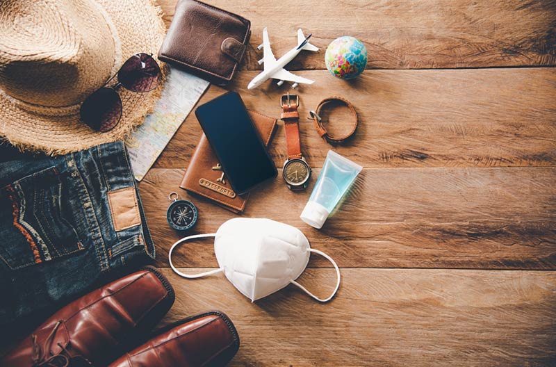 A collection of travel packing-worthy items against a brown wood background: straw hat, sunglasses, wallet, small glove, compass, mask, and visas.