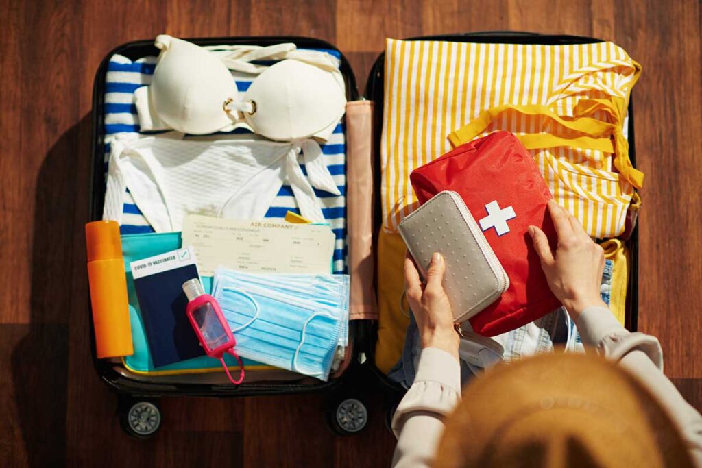 Top-down view of a person packing a suitcase for a trip, placing a first-aid kit on top of a shirt in open luggage.