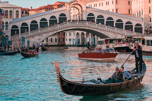 A gondola carrying people glides through the Grand Canal in Venice, with the Rialto Bridge in the background. Other boats can be seen on the water, and historic buildings line the canal under a clear sky.