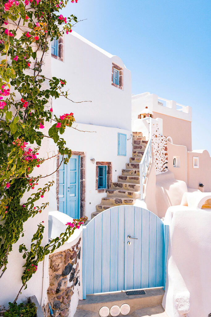 A picturesque Mediterranean house with whitewashed walls, a light blue wooden gate, and a staircase. Pink bougainvillea flowers climb the facade under a clear blue sky.