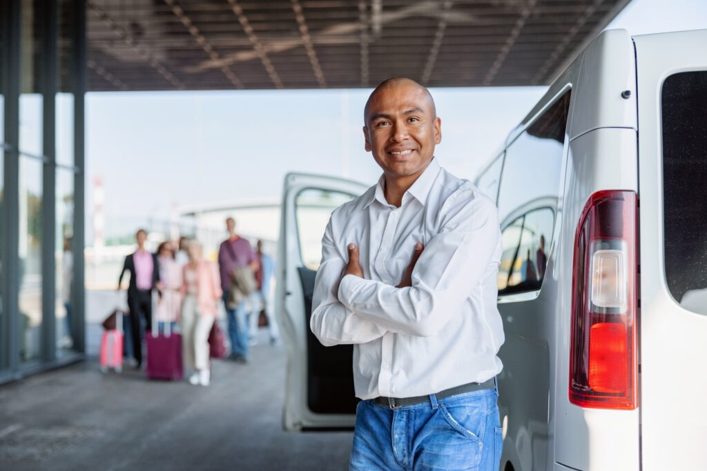 A man in a white shirt and jeans stands confidently with arms crossed in front of a white van at a transportation hub. In the background, a group of people with luggage are approaching.